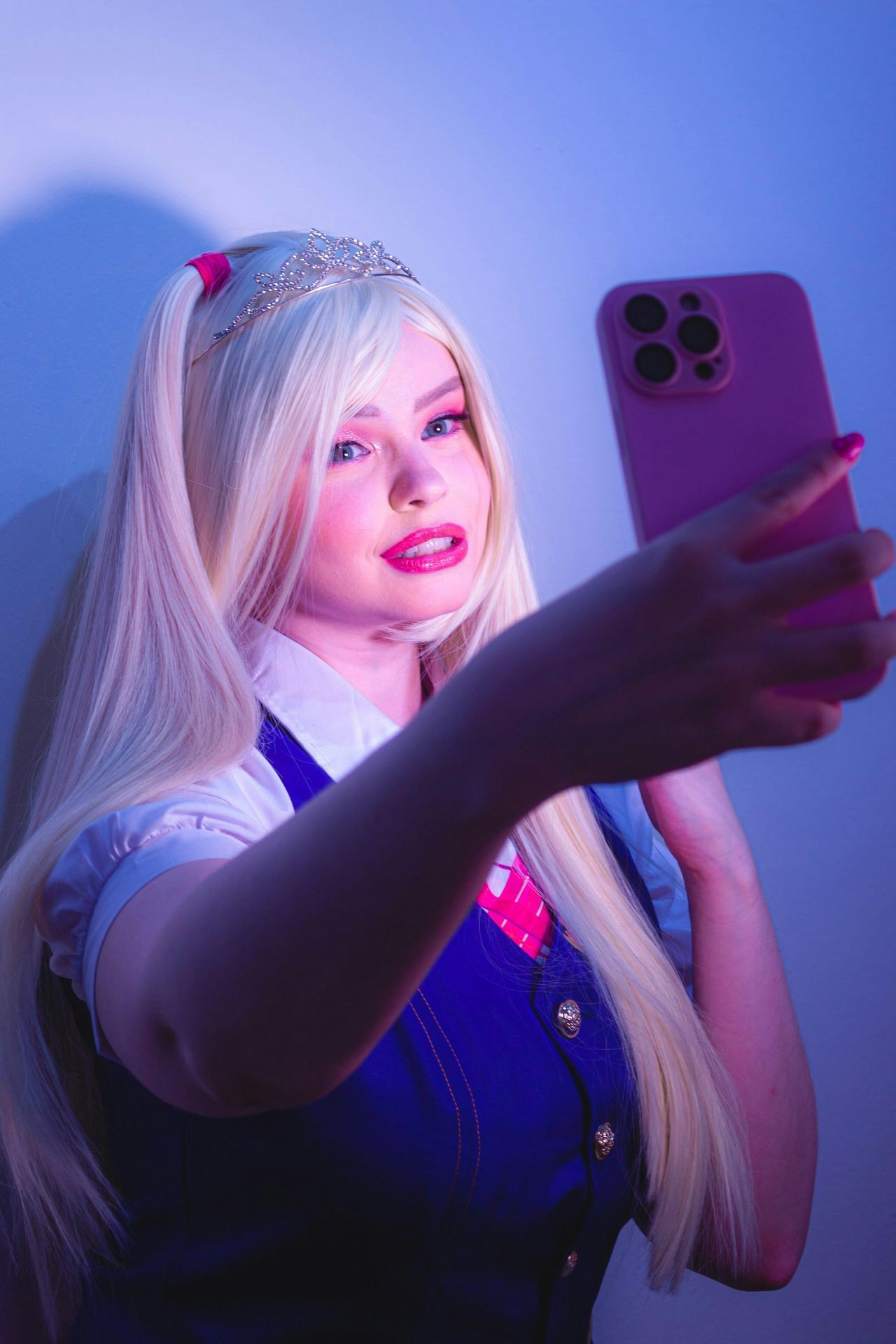 Person with long blonde hair wearing a silver tiara and school uniform, holding a pink smartphone.